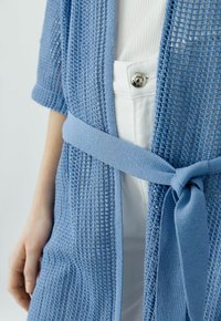 Hellblauer Strickcardigan mit einem taillierten Gürtel, der ein texturiertes, offenes Webmuster und kurze Ärmel aufweist, über einem weißen Top mit Knopf.