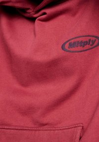 Czerwona bluza z kapturem wykonana z bawełnianej mieszanki tkanin, z przednią kieszenią oraz czarnym, zaokrąglonym logo na piersi. Gładka tekstura.