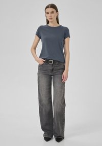 Dunkelgraues T-Shirt mit kurzen Ärmeln, kombiniert mit hochtaillierten schwarzen Jeans. Das Model trägt einen schwarzen Gürtel mit einer metallischen Schnalle.