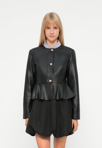 Jeune femme aux cheveux blonds raides portant une veste peplum noire avec des boutons dorés sur une chemise à col rayé et un short noir.