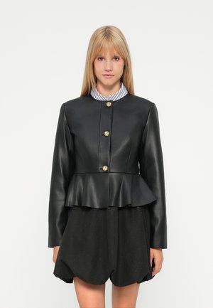 Jeune femme aux cheveux blonds raides portant une veste peplum noire avec des boutons dorés sur une chemise à col rayé et un short noir.