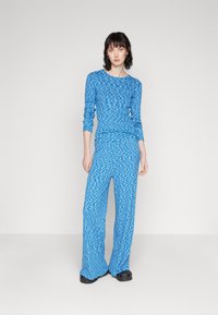 Blue patterned long-sleeve top and matching wide-leg pants in a textured, wave design. Material appears soft and fluid. Black footwear.