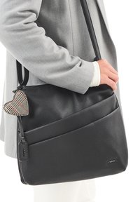 Bolso de hombro de cuero texturizado negro con bolsillos con cremallera y un charm decorativo en forma de corazón de tela. El bolso tiene una larga correa ajustable.