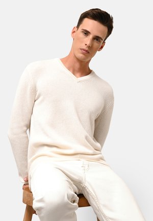 Suéter color beige claro con escote en forma de V, confeccionado en tela de punto texturizada, con mangas largas y puños acanalados, combinado con pantalones color crema, sentado en un taburete de madera.
