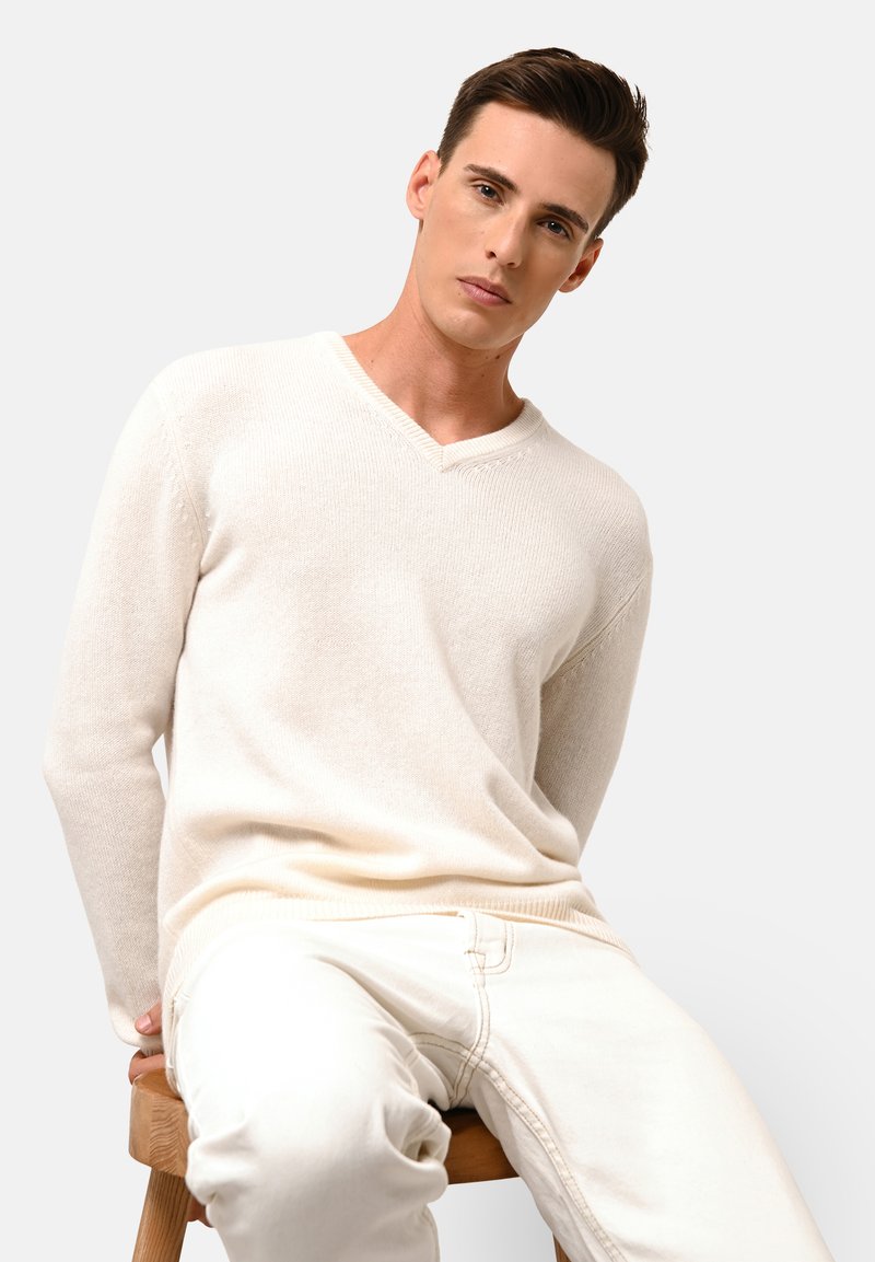 Hellbeige V-Ausschnitt-Pullover aus strukturiertem Strickstoff, mit langen Ärmeln und gerippten Bündchen, getragen mit cremefarbenen Hosen, sitzend auf einem Holzschemel.