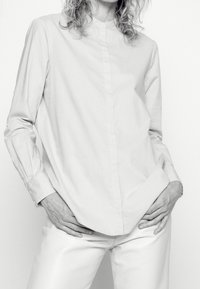 Ljust grå skjorta med ståkrage, långa ärmar och rak skärning. Slät tyg med minimalt synliga sömmar.