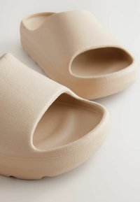 Sandales ouvertes beiges avec une surface texturée, un design à bout ouvert et une semelle lisse et arrondie, présentant une forme minimaliste et moderne.