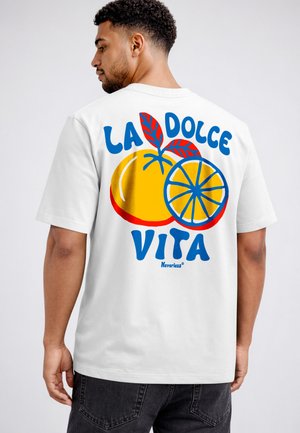 Man wearing white t-shirt with colorful orange fruit and blue text "LA DOLCE VITA" on the back, standing against a plain background.