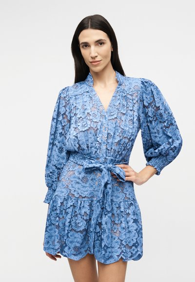 Woman with long dark hair wearing a blue floral lace dress with puffed sleeves and a tied waist belt, standing against a plain background.