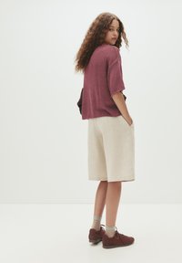 Bordeaux-rotes kurzärmliges Oberteil, beiges knielanges Shorts und burgunderrote Schuhe. Die Haare sind lockig; die Hände in den Taschen. Minimalistischer Hintergrund.