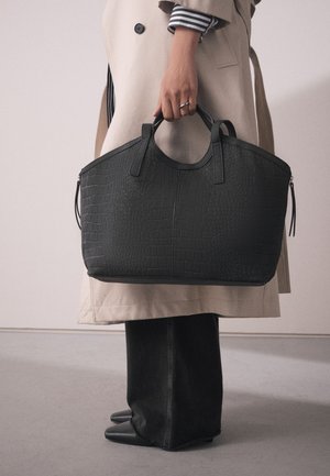 Black crocodile texture leather tote bag with two sturdy handles, a zip closure, and a structured silhouette. Worn with a beige coat and dark pants.