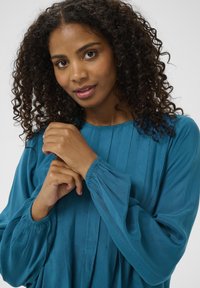 Teal blouse met lange mouwen, voorzien van een geplooid ontwerp en elastische manchet, gemaakt van zachte stof. Model houdt haar handen bij de halslijn.