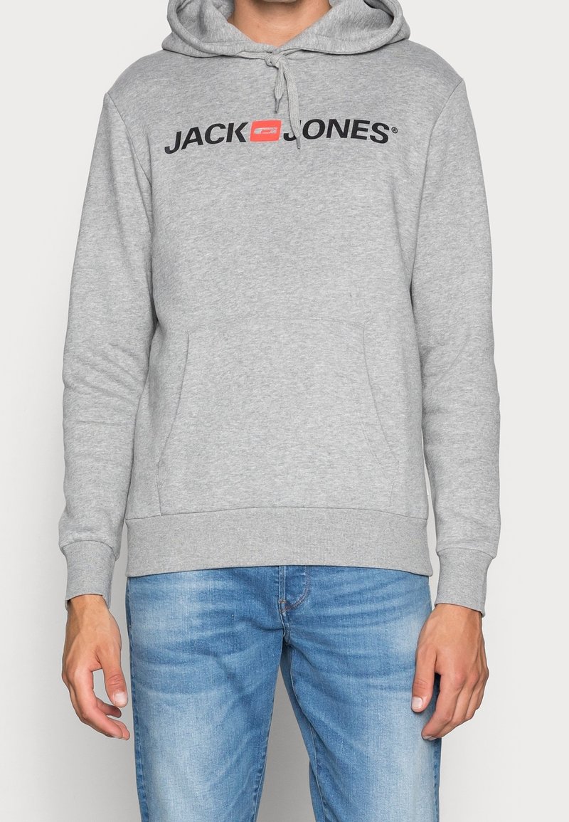 Sudadera gris con bolsillo canguro, capucha con cordón y un logo negro en el frente. Combinada con jeans de mezclilla azul.