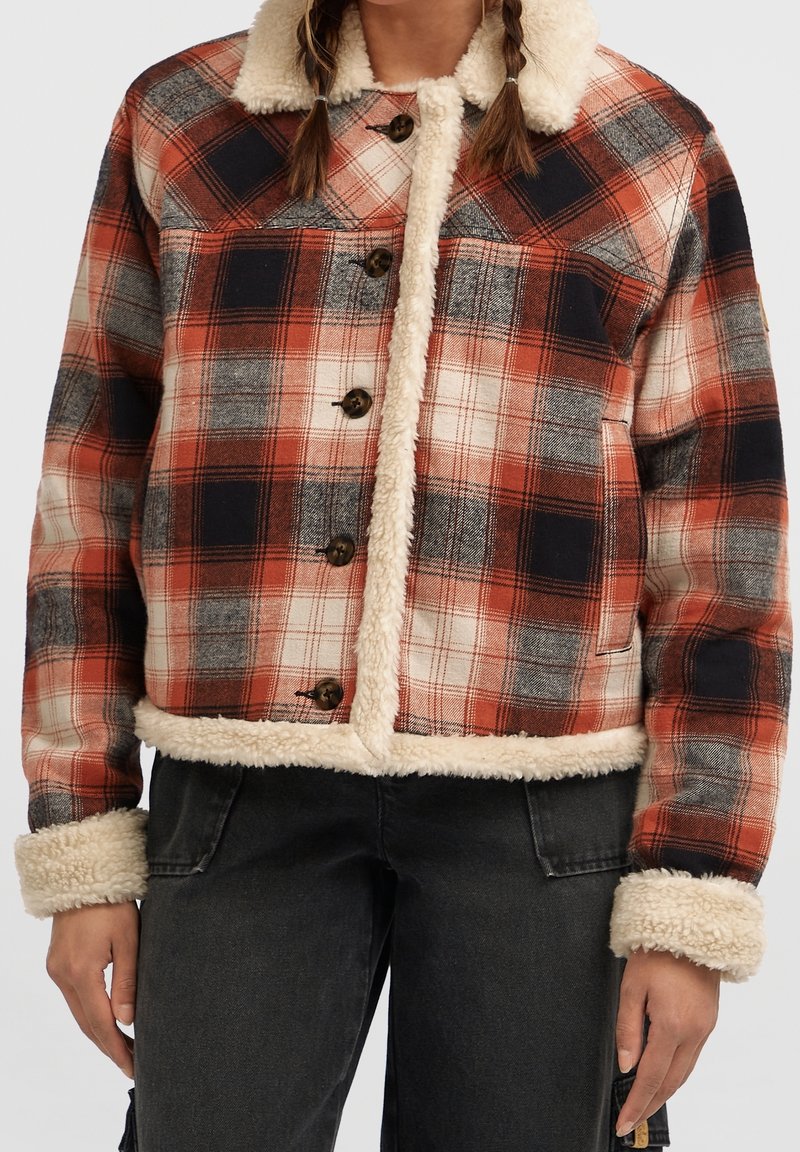 Femme portant une veste à carreaux rouge, noire et beige avec col, poignets et ourlet en shearling, associée à un pantalon noir.