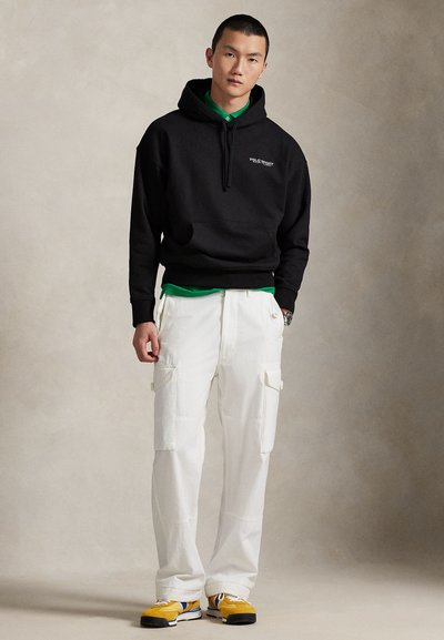 Juodas džemperis su gaubtu, priekyje kišenė ir raišteliai, dėvimas ant žalios spalvos marškinėlių. Baltos kargo kelnės su šoninėmis kišenėmis ir geltonais sportbačiais.