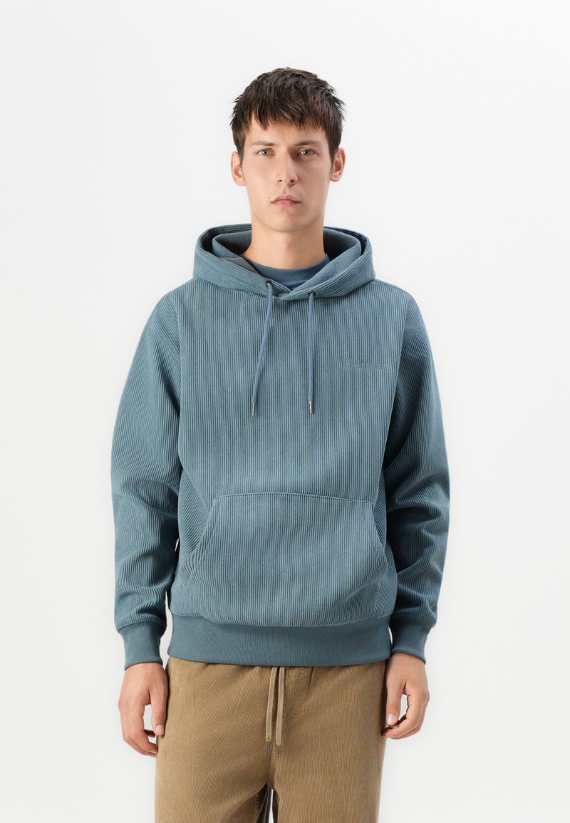 Sudadera con capucha de tela acanalada en verde azulado, con un bolsillo tipo canguro en la parte delantera y cordones ajustables, combinada con pantalones beige con cordones.