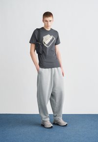 Γκρι μπλουζάκι Nike με λογότυπο, μικρή μαύρη τσάντα χιαστί, γκρι άνετο παντελόνι φόρμας και γκρι αθλητικά παπούτσια μπροστά σε απλό φόντο.