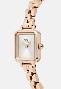 Rozā zelta taisnstūra pulkstenis ar metāla siksniņu, baltu saulespuķes stila ciparnīcu un kroni sānos. Uz ciparnīcas redzams "Daniel Wellington" logo.