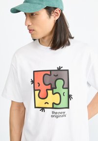 Weißes T-Shirt mit einem bunten Puzzle-Teil-Grafik in Rot, Gelb, Grün und Grau, versehen mit cartoonhaften Gesichtsausdrücken und Details.