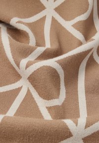 Béžová pletená tkanina s bielymi prekrývajúcimi sa zakrivenými a rovnými líniami vytvárajúcimi geometrický vzor, zobrazená s vráskami a detailmi textúry.