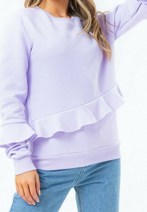 Kvinde iført lyse lilla sweatshirt med flæsedetalje og blå jeans, stående med en arm løftet.