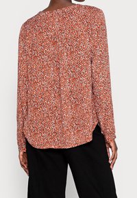 Blouse à manches longues de couleur orange avec un motif abstrait noir et blanc. Présente un ourlet arrondi et un tissu doux et léger.