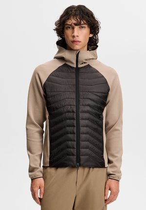 Młody mężczyzna z kręconymi włosami, ubrany w beżowo-czarną bluzę z kapturem na zamek, stojący na jednolitym tle.