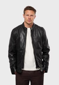 Black leather jacket with a stand collar, zip closure, and smooth texture. Worn over a white crew-neck shirt, featuring subtle stitching details.