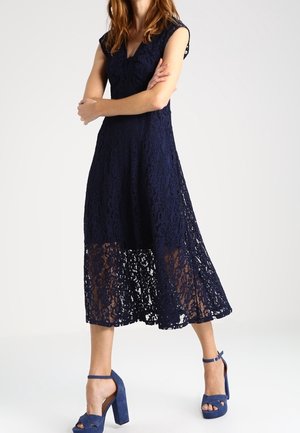 Femme portant une robe midi en dentelle bleu marine sans manches et des sandales à talons assorties, debout les bras croisés.