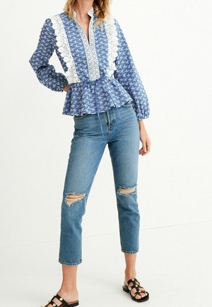 Kvinde iført blå mønstret bluse med hvide blonde detaljer, slidte cropped jeans og sorte sandaler med nitter, stående mod en hvid baggrund.