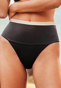 Femme portant un bas de bikini noir taille haute avec une ceinture grise, debout en plein air les bras croisés à la taille.