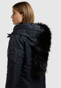 Cappotto imbottito blu navy con collo in pelliccia sintetica nera. Presenta cerniere e cappuccio con coulisse. Tasca laterale con dettaglio elasticizzato.