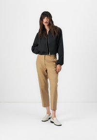 Giacca bomber nera corta, pantaloni beige a sigaretta e scarpe slip-on bianche. Tessuto liscio e linee pulite con dettagli minimi.