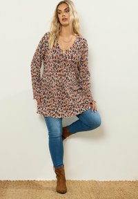 Blommig tunika med V-ringning, långa ärmar och volang nederkant; har nyanser av brunt och rött på en krämfärgad bakgrund, kombinerad med blå jeans.