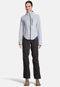 Veste imperméable gris clair avec un col haut, une poche poitrine zippée et des accents noirs, associée à un pantalon noir et des bottes marron clair.