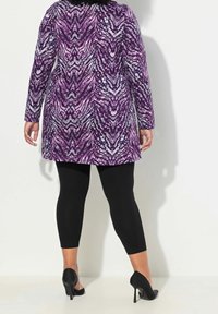 Tunique à motifs violets et gris à manches longues, associée à un legging noir et des chaussures à talons, vue de dos.