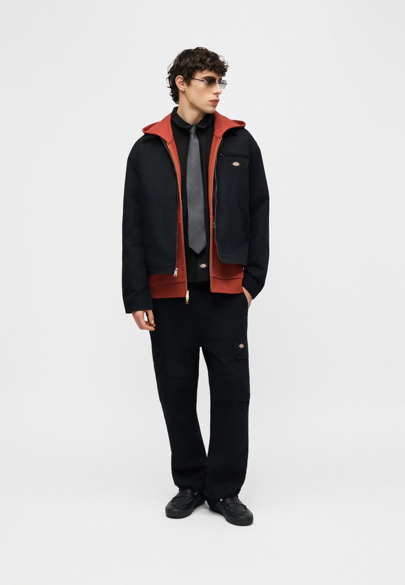 Junger Mann, der schwarze und rote Reißverschlussjacken, schwarze Cargohosen, graue Krawatte, Sonnenbrille und schwarze Schuhe trägt, steht mit entspannten Händen.