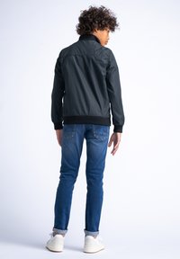 Zwarte windbreaker met een ritssluiting aan de voorkant, geribbelde zoom en boorden, gecombineerd met blauwe jeans en witte sneakers, gezien vanuit de achterkant.