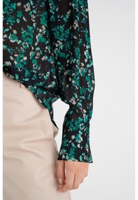 Blusa nera con motivo floreale verde e crema, tessuto trasparente, maniche lunghe e vestibilità rilassata. Indossata con pantaloni chiari.