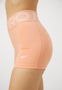 Șorturi Nike Pro de culoare piersică deschisă, realizate dintr-un material elastic. Prezintă un brâu lat, cu text și un logo subtil pe coapsă.