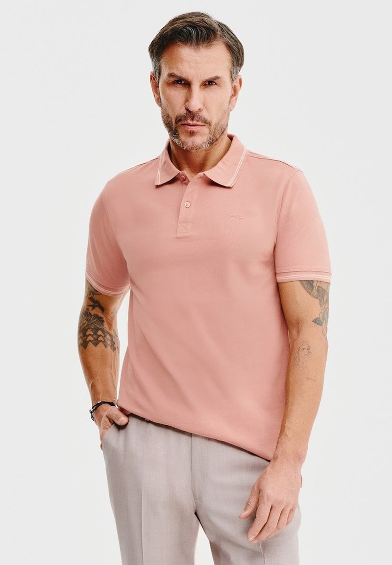 Jasnoróżowa koszulka polo z trzema guzikami, kontrastowym kołnierzykiem i krótkimi rękawami. Wykonana z miękkiego materiału, z delikatnym logo, noszona z szarymi spodniami.
