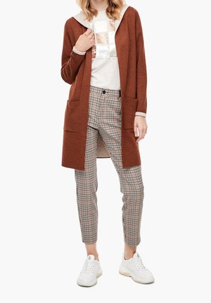 Vrouw draagt een roestkleurige open cardigan, wit grafisch T-shirt, geruite smalle broek en witte sneakers, staand tegen een witte achtergrond.