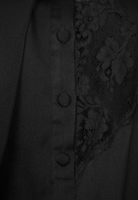Blusa nera con pannello in pizzo sul lato sinistro, bottoni tondi lungo la parte frontale e una consistenza liscia e morbida in tutto il capo.