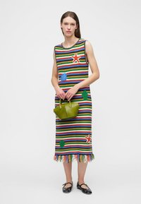 Frau trägt ärmelloses, mehrfarbig gestreiftes Kleid mit Meerestier-Motiven, hält kleine grüne Handtasche, schwarze Ballerinas, steht vor weißem Hintergrund.