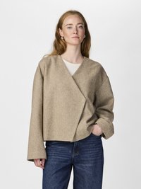 Jacheta bej, stil wrap, dintr-un material texturat cu mâneci largi. Asortată cu blugi de culoare albastru închis, designul prezintă o silueta modernă, relaxată.