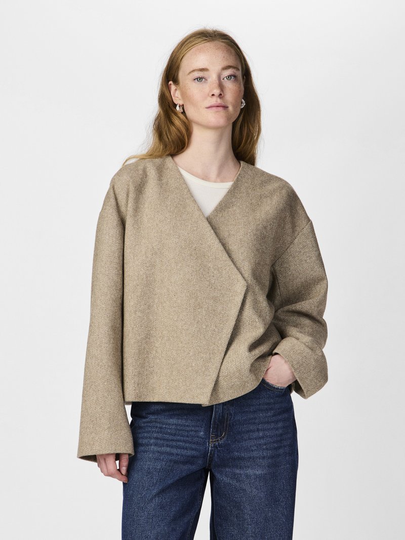 Jacheta bej, stil wrap, dintr-un material texturat cu mâneci largi. Asortată cu blugi de culoare albastru închis, designul prezintă o silueta modernă, relaxată.