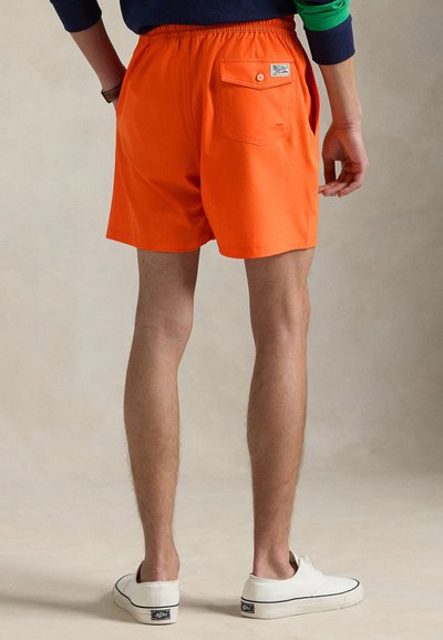 Πορτοκαλί σορτς μέχρι το μέσο του μηρού με ελαστική μέση, επίπεδη τσέπη στο πίσω μέρος και απαλή υφή. Φοριούνται με λευκά αθλητικά παπούτσια και φόντο ουδέτερου χρώματος.