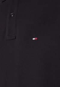 Schwarzes Poloshirt aus strukturiertem Stoff, mit einem Drei-Knopf-Verschluss und einem kleinen gestickten Logo in Rot, Weiß und Blau.