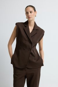 Frau in einem ärmellosen, dunkelbraunen, doppeltknöpfigen Blazer und passenden Hosen, mit goldenen Creolen, posierend vor einem weißen Hintergrund.