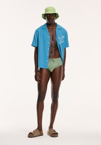 Srajca s kratkimi rokavi modre barve z gumbi, zelene kopalne hlače in zelena čelada. Rjave sandale. Gladek material, sproščen kroj, preprost dizajn.